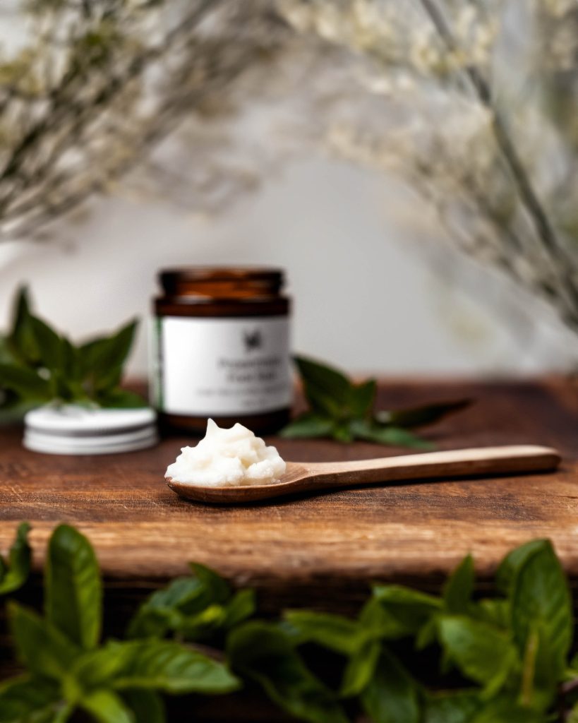 A wooden spoon with peppermint foot rub. In the background is a blurred jar and peppermint leaves